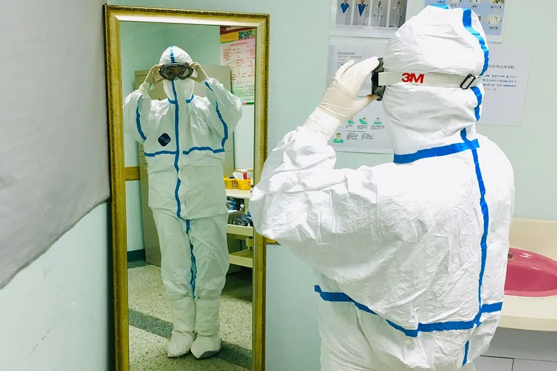 Doctor in protective scrubs looks at self in mirror.