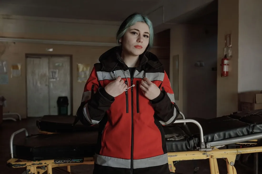 White woman with blue hair and red jacket stands in front of gurney.