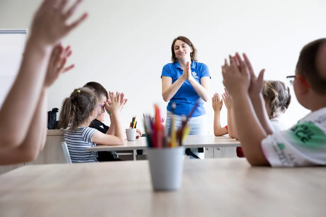 White woman in blue shirt claps along with children in a classroom