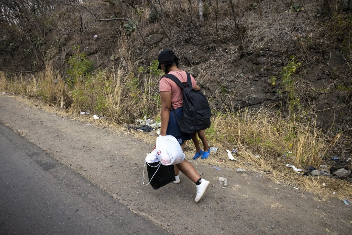 A woman carrying a child in one arm and bags in another, walks along a road