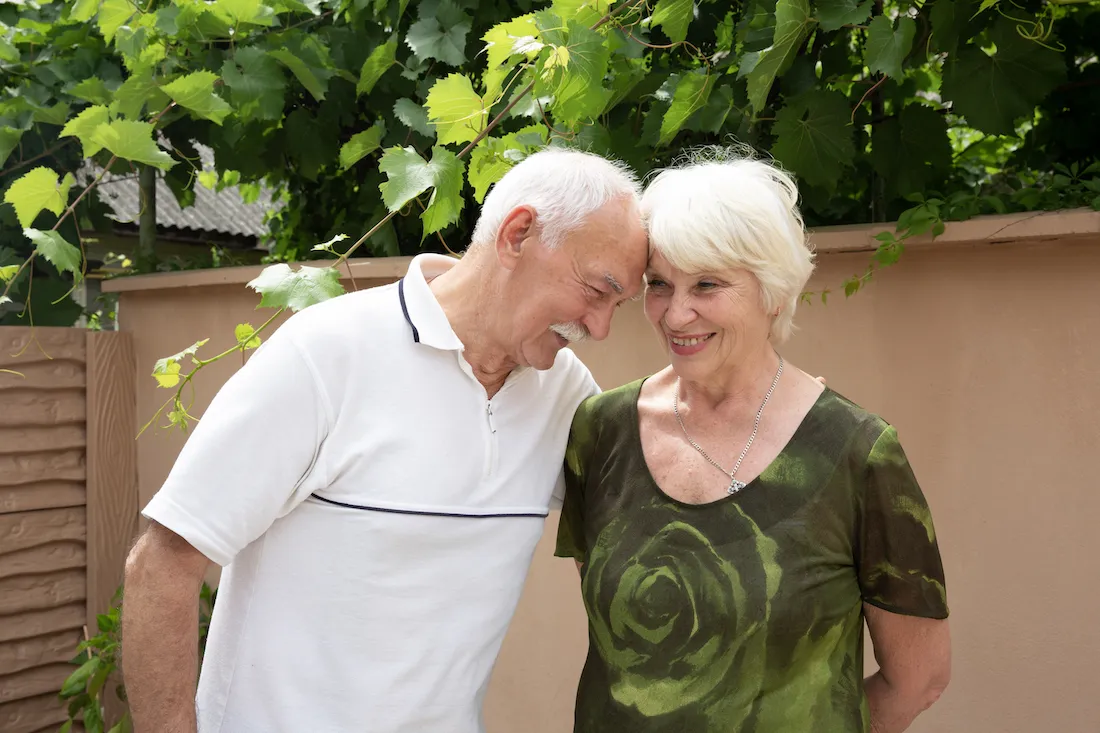 An elderly white man and woman smile and touch heads in affection