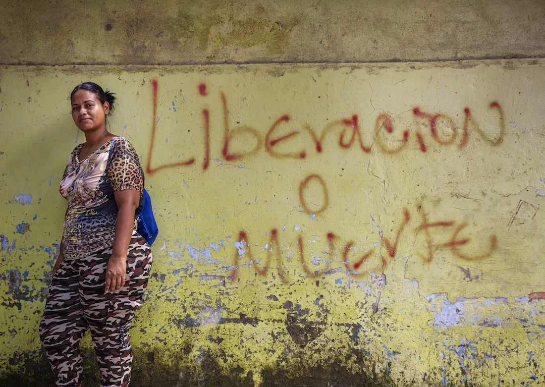 a woman in pink camo and leopard print stands in front of a yellow wall that reads in Spanish "Liberation or Death"